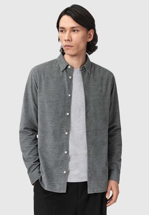 Junger Mann mit dunklen Haaren trägt ein graues Hemd über einem hellgrauen T-Shirt und steht mit den Händen in den Taschen vor einem schlichten Hintergrund.