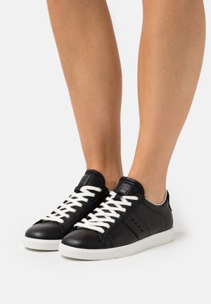 Benen met zwarte lage sneakers met witte veters en witte zolen, getoond tegen een effen lichte achtergrond.