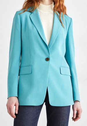 Lyseblå blazer med notch-lapper, enkelt knaplukning og to forlommer. Båret over en cremefarvet rullekrave og mørke jeans.