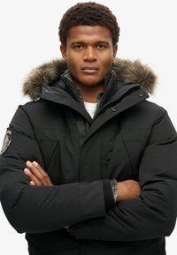Chaqueta acolchada negra con capucha de piel sintética, múltiples bolsillos y cierre de cremallera, superpuesta sobre una camisa de cuadros.