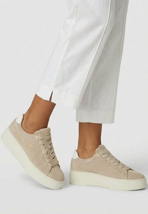 Persona que lleva zapatillas de plataforma de ante beige con suelas blancas gruesas y pantalones blancos recortados con aberturas laterales, de pie sobre una superficie blanca.