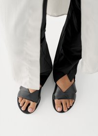 Sandales en cuir noir avec des brides croisées, semelle plate et bout ouvert. Les pieds sont positionnés sous un vêtement blanc, sur un fond uni.