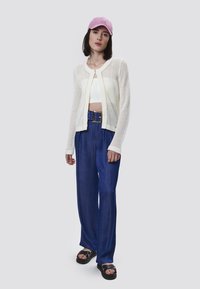 Cárdigan de malla blanco sobre un top corto blanco, combinado con pantalones anchos azul y una gorra rosa. Sandalias de plataforma negras completan el atuendo.