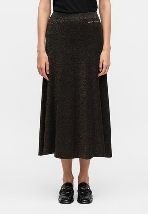 Kvinde iført en sort glitrende midi-nederdel med Armani Exchange-logo og sorte loafers, stående mod en hvid baggrund.