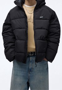Giacca a vento nera con cappuccio, caratterizzata da una chiusura con zip, texture trapuntata e un piccolo logo. Abbinata a una camicia beige e pantaloni in denim.