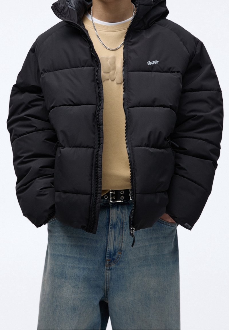 Giacca a vento nera con cappuccio, caratterizzata da una chiusura con zip, texture trapuntata e un piccolo logo. Abbinata a una camicia beige e pantaloni in denim.
