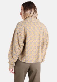 Chemise en flanelle jaune avec un motif de tartan géométrique en gris et rose. Elle présente un poignet à boutons et un patch logo sur l'épaule gauche.