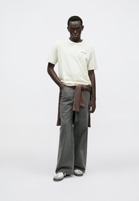 Ljusgul t-shirt med logotyp, kombinerad med vida grå jeans och en brun tröja knuten i midjan; bäraren har robusta sneakers, neutral bakgrund.