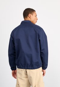 Jachetă de vânt bleu marin cu guler și manșete elastice, are un design simplu și o croială lejeră, purtată peste pantaloni bej.