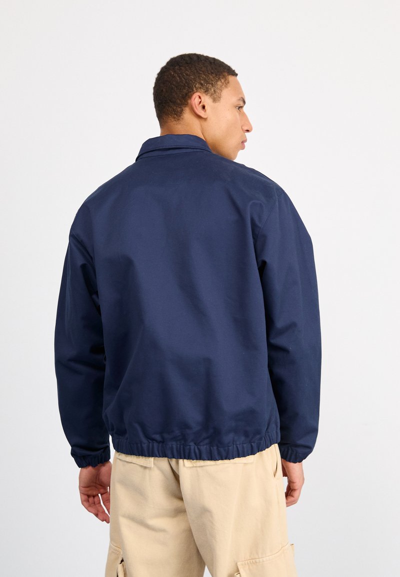 Veste coupe-vent bleu marine avec un col, des poignets élastiques, dotée d'un design simple et d'une coupe décontractée, portée sur un pantalon beige.