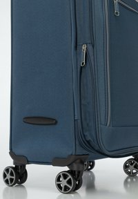 Valise bleue texturée avec plusieurs compartiments zippés, roues noires et poignée moulée sur le côté pour une manipulation facile.