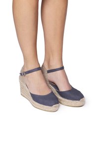 Espadrilky na klínu s námořnickým textilním svrškem, jutovou mezipodešví a pásem kolem kotníku. Zaoblená špička a texturované povrchové detaily.