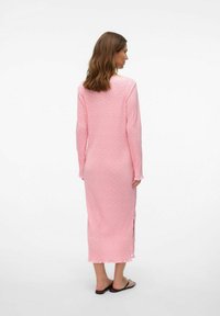 Lange, roze bloemenjurk van zachte stof, met lange mouwen, zijsplitten en een geschulpte zoom, gedragen met zwarte sandalen.