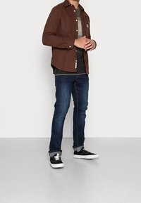 Camicia marrone a maniche lunghe con bottoni, jeans in denim e sneakers nere. La camicia ha una tasca sul petto e uno strato sottostante verde scuro texturizzato.
