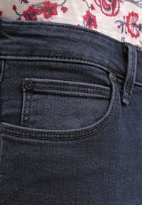 Jeans de mezclilla oscura con una vista en primer plano de la cintura, que presenta un bolsillo pequeño y detalles de costura sutiles. Tela texturizada, diseño de tiro medio.