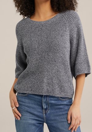 Kvinde iført en grå strikket sweater med albuelange ærmer og blå jeans, poser med den ene hånd hvilende på hoften.