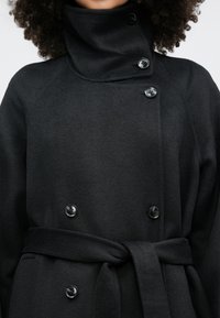 Kvinde iført en sort dobbeltradet frakke med store knapper, høj krave og et bånd bundet om taljen, der viser den øverste del af overkroppen.