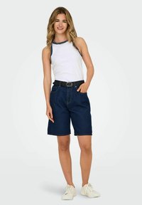 Shorts in denim con vita alta, di colore blu scuro, con orlo cucito; abbinati a un top senza maniche bianco con dettagli blu navy e una cintura nera.