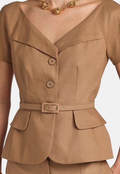 Woman wearing a tan short-sleeve belted blazer with buttons, flap pockets, and a gold knot-style necklace.