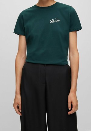 Osoba ubrana w ciemnozieloną koszulkę z krótkim rękawem z białym napisem "Be a Boss" na piersi oraz luźne czarne spodnie na jednolitym tle.