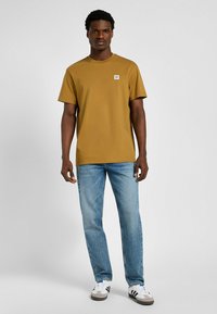 Senfgelbes kurzärmeliges T-Shirt mit rundem Ausschnitt, das mit einem kleinen weißen Logo-Patch versehen ist. Kombiniert mit hellblauen Jeans und Sneakers.