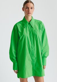 Chemise verte surdimensionnée avec un col pointu, des manches longues, une poche poitrine et une texture lisse et brillante.
