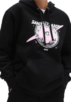Personne portant un sweat à capuche Vans noir avec une tête de chat rose dessin animé, un grand œil vert, une planète annelée et un texte blanc à l'avant. Mains dans les poches.