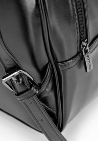 Bolso de mano de cuero negro con una textura suave, correa ajustable, herrajes metálicos y un cierre de cremallera con un tirador de cremallera de marca.