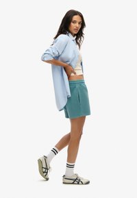 Camisa de botones azul claro, camiseta sin mangas crema, pantalones cortos verde azulado, calcetines blancos con rayas negras y zapatillas beige con acentos en azul marino.