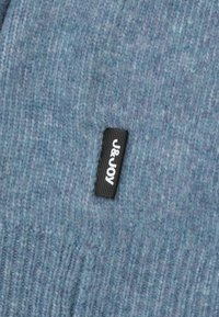 Gebreide blauwe muts met een zachte textuur, met een zwart label met de tekst "J&JOY" bevestigd aan de zijkant.