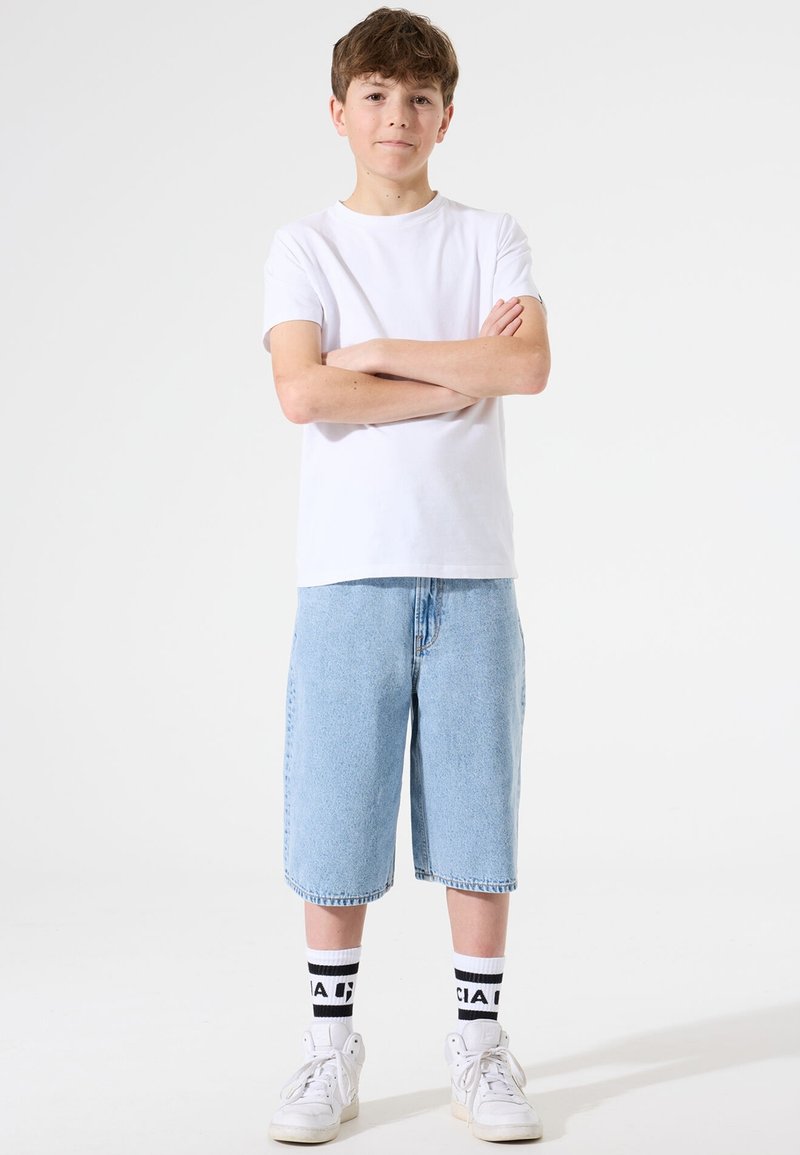 T-shirt en coton blanc associé à un short en denim bleu clair, des baskets blanches et des chaussettes noires et blanches avec un logo. Tenue décontractée simple.