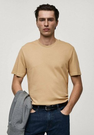 Beige cotton t-shirt with short sleeves, round neckline, worn with a gray hoodie over the arm. Paired with blue denim jeans and a black belt.
