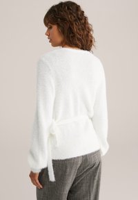 Kortsirged juuksed ja valge sokiülikond, millel on külje sidumisse detail ja hallist ruudulistest pükstest, selg ees neutraalse taustaga.