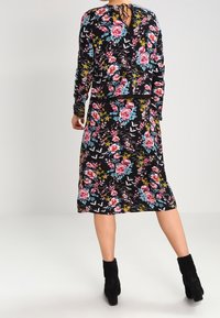 Abito floreale con maniche lunghe, fondo nero, fiori colorati in rosa, blu e giallo. Presenta un dettaglio con laccio sul retro e un design fino al ginocchio.