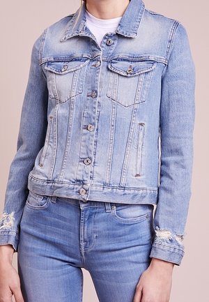Lichtblauw spijkerjasje met knoopsluiting en borstzakken, gedragen over een witte top, gecombineerd met bijpassende lichtblauwe spijkerbroeken.