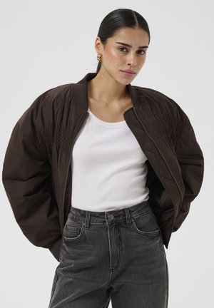 Femme aux cheveux foncés attachés en arrière, portant des boucles d'oreilles créoles dorées, une veste bomber marron foncé sur un débardeur blanc et un jean noir taille haute.