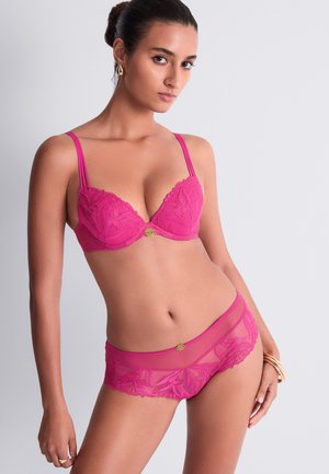 Femme portant un ensemble assorti de soutien-gorge et culotte en dentelle rose avec des accents décoratifs dorés, debout devant un fond clair uni.