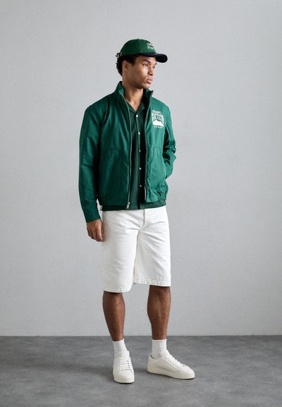 Veste légère verte avec deux poches avant, associée à une chemise boutonnée vert foncé et à un short en denim blanc. Baskets blanches et casquette assortie.
