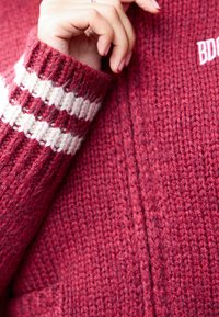 Hand with red nail polish touches collar of thick red knit sweater with white striped cuff and partial white embroidered letters.