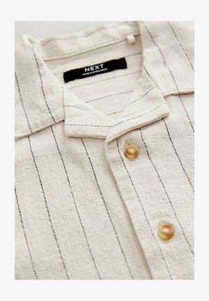 Bežová košeľa s golierom vyrobená z textúrovanej tkaniny s vertikálnymi čiernymi pruhmi, s gombíkovým zapínaním a štítkom na golieri.