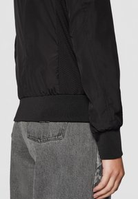 Gros plan d'une personne portant une veste noire avec des poignets et un ourlet côtelés, et un jean en denim gris avec une poche arrière visible.