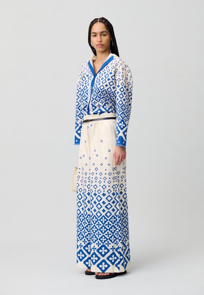 Vestido largo con fondo blanco y patrones geométricos azules, con un frente abotonado y puños azules, acompañado de un bolso beige.