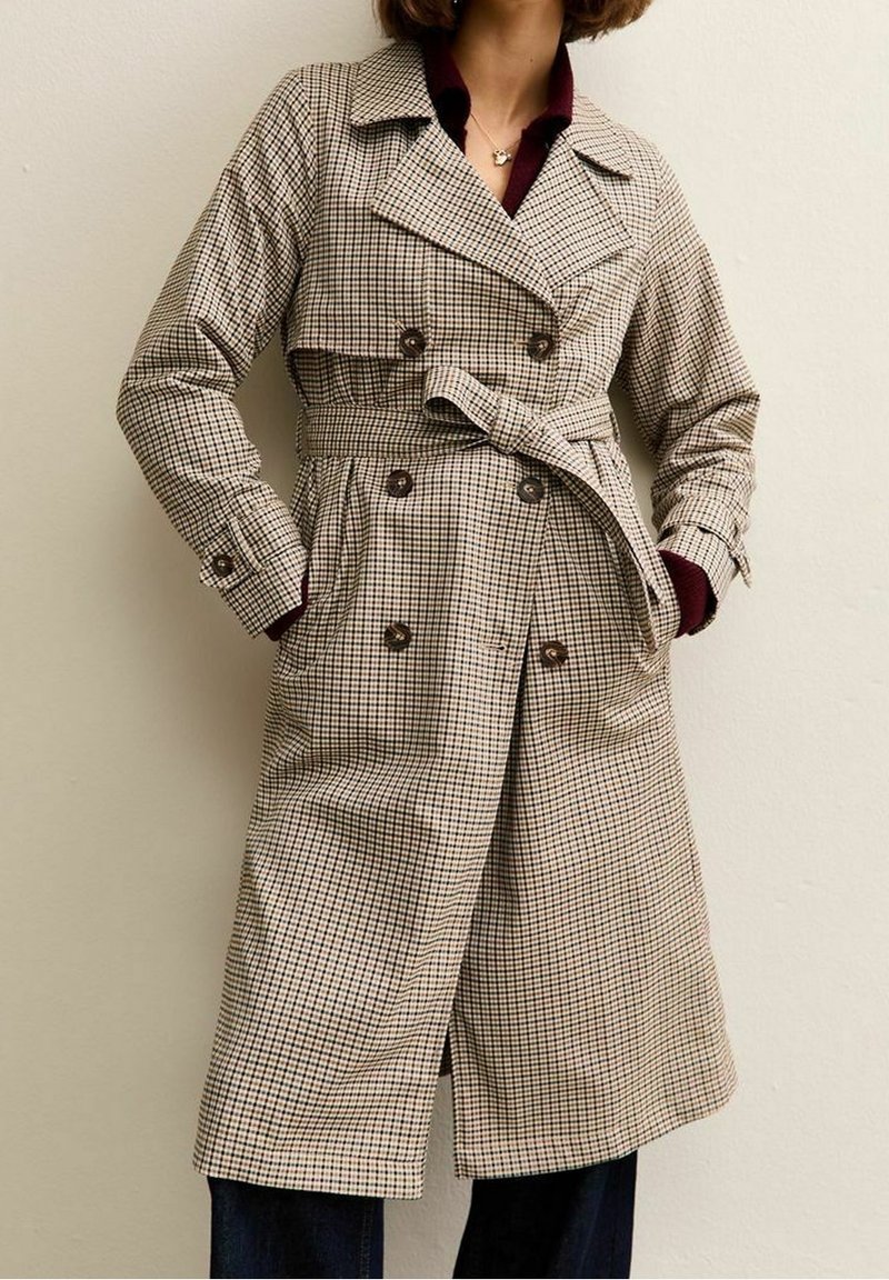 Femme portant un trench-coat beige et marron à carreaux, double boutonnage, avec ceinture et poches, assorti à un pantalon foncé et une chemise rouge foncé en dessous.