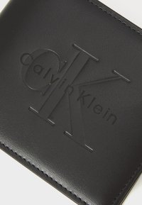 Portefeuille en cuir noir avec texte et logo "Calvin Klein" en relief. Texture lisse avec des coutures le long des bords. Forme rectangulaire.