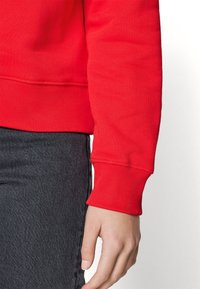 Rode sweatshirt met geribbelde manchetten, zachte textuur en een iets losser vallende pasvorm, gecombineerd met donkergrijze spijkerbroek. Close-up van de arm en taille.