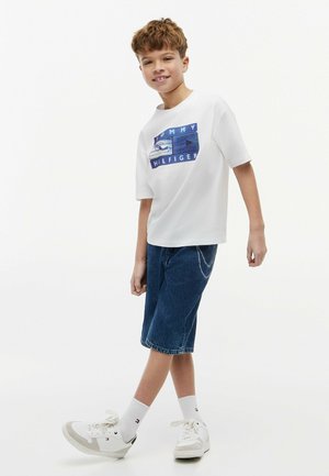 Boy smiling and walking, wearing a white Tommy Hilfiger t-shirt, blue denim shorts, white socks, and white sneakers.