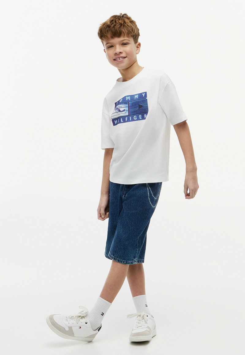 Garçon souriant et marchant, portant un t-shirt blanc Tommy Hilfiger, un short en jean bleu, des chaussettes blanches et des baskets blanches.