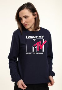 Marinblå sweatshirt med en framtida grafik med texten "JAG VILL HA MIN MTV" i vit och rosa, rund halsringning och långa ärmar.