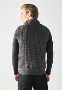 Gilet en tricot gris avec un col haut, motif texturé et ourlet côtelé. Porté par-dessus une chemise à manches longues noire, vu de dos.
