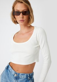 Weißes, geripptes, langärmliges Crop-Top mit breitem Ausschnitt, kombiniert mit hochgeschnittenen blauen Jeansshorts. Der Hintergrund ist schlicht weiß.